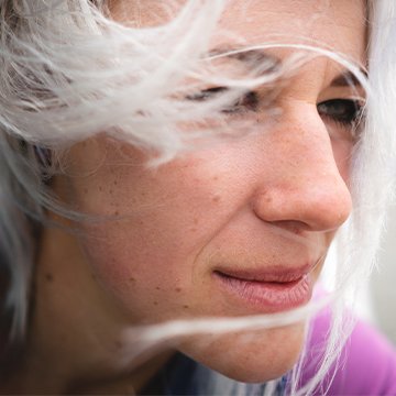 Close-up of a person with white hair covering part of their face, wearing a purple garment.
