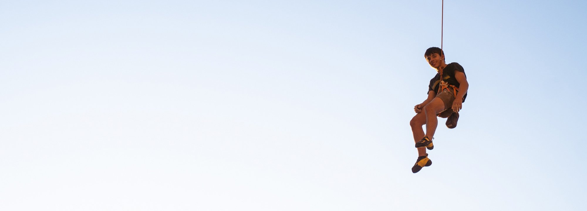 A person wearing climbing gear and a harness is suspended in mid-air against a clear blue sky, holding onto a rope and looking down.
