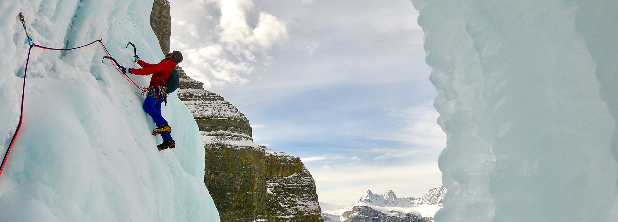 A climber wearing winter gear ascends a vertical, icy wall using ice axes and crampons, secured by ropes, with snow-covered mountains and a cloudy sky visible in the background.