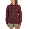 A woman wears the La Sportiva Andes Long Sleeve Shirt Women's in red and dark gray plaid, paired with khaki shorts. The shirt, made from recycled polyester, features visible La Sportiva branding on the chest and sleeve.