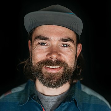 Man with a beard and cap smiling against a black background