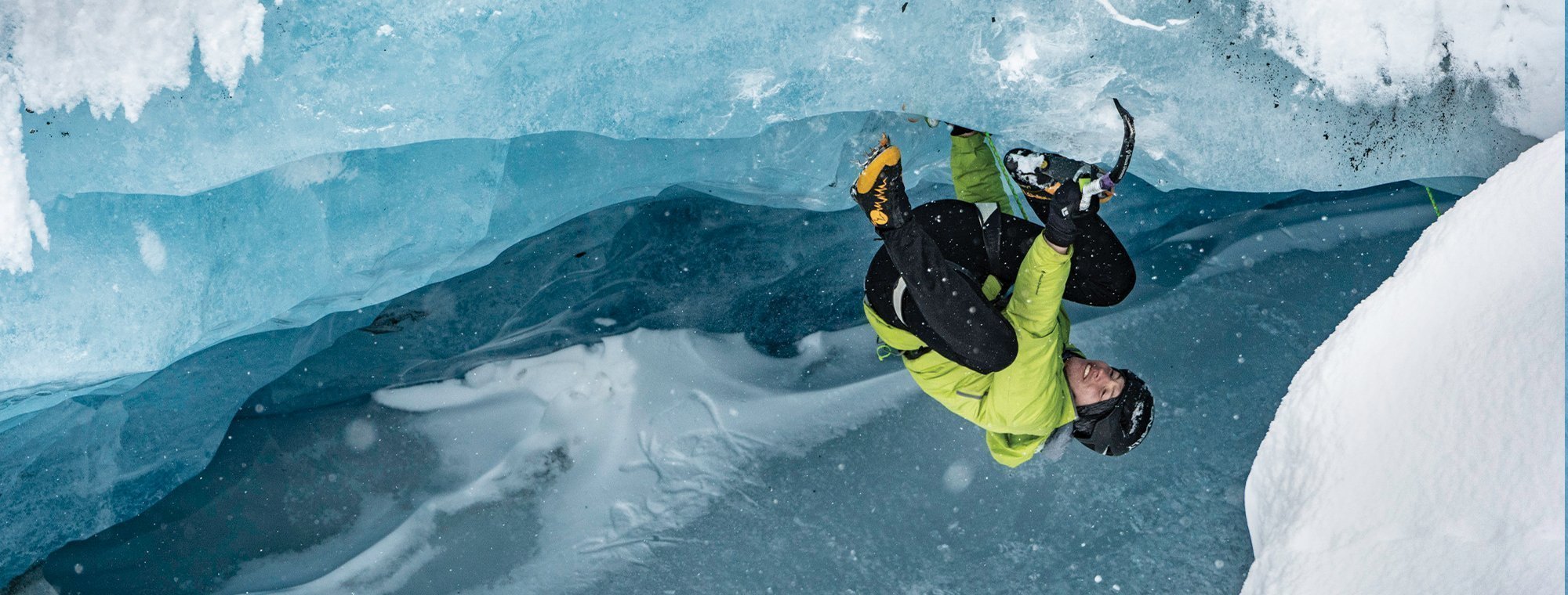 Person in a yellow jacket climbing ice with tools against a blue ice wall.