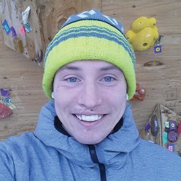 Person wearing a colorful knit hat and blue jacket in front of a wooden wall with decorations.