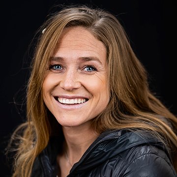 Woman with long blonde hair wearing a black jacket against a black background